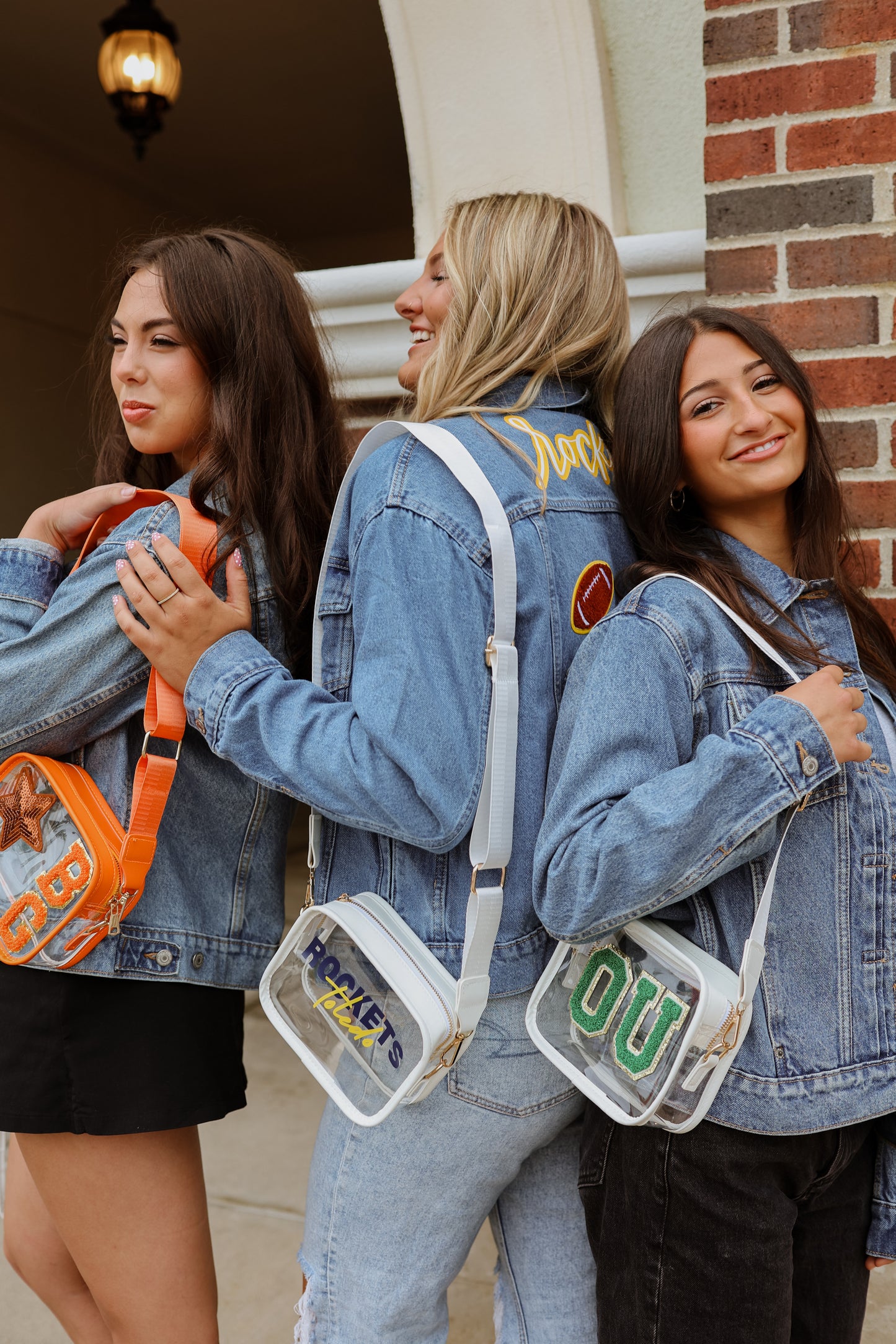 Three women wearing denim jackets with unique patches, standing in front of a brick building holding their clear stadium purses