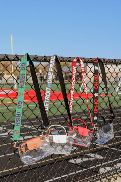 Collection of clear purses with sports team-themed beaded straps on a chain-link fence with a football field background.
