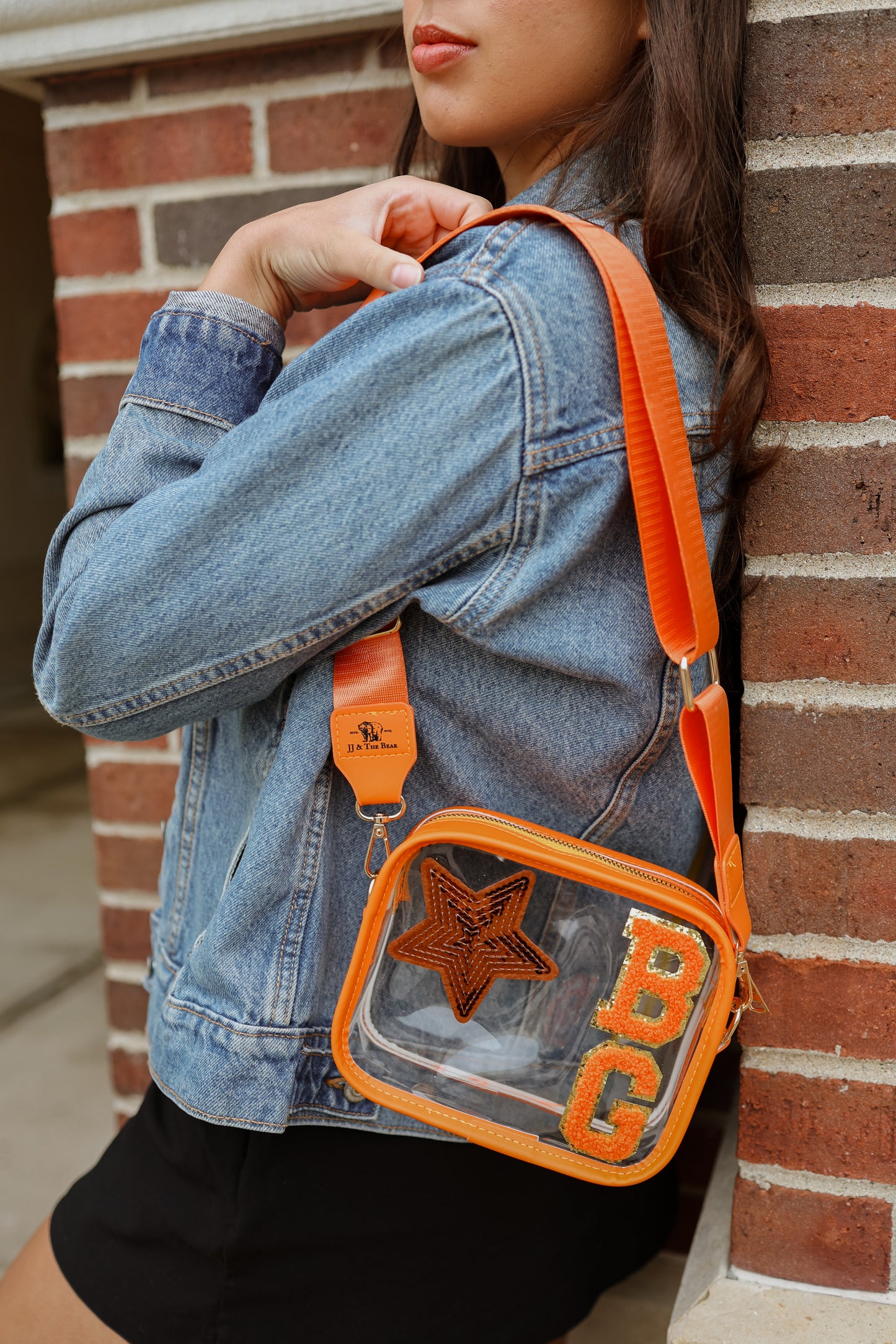 Person wearing a denim jacket with an orange crossbody bag featuring a star and 'B G' initials against a brick wall.