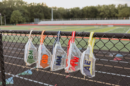 Fanny pack bags with various designs and text hanging on a chain-link fence with a sports field background