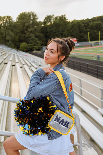 Woman in a denim jacket with a 'HAIL' bag and pom-poms sitting on bleachers at a sports field.