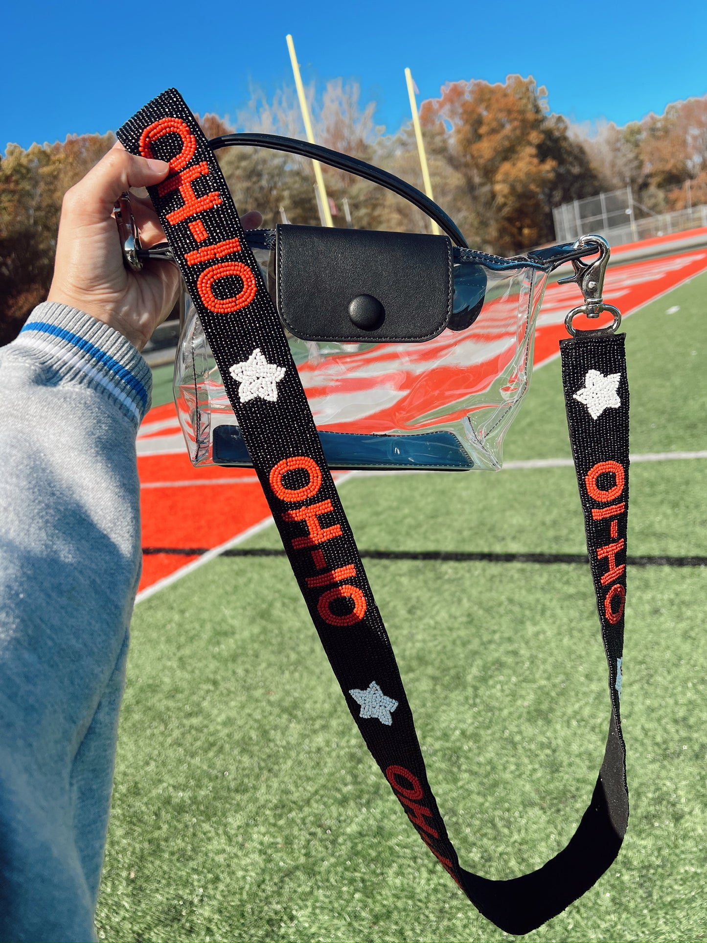 Clear purse with black handle with 'OHIO' beaded strap held over a sports field.