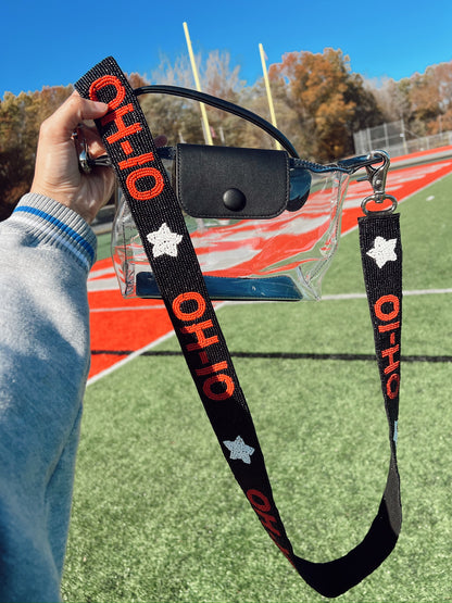 Clear purse with black handle with 'OHIO' beaded strap held over a sports field.