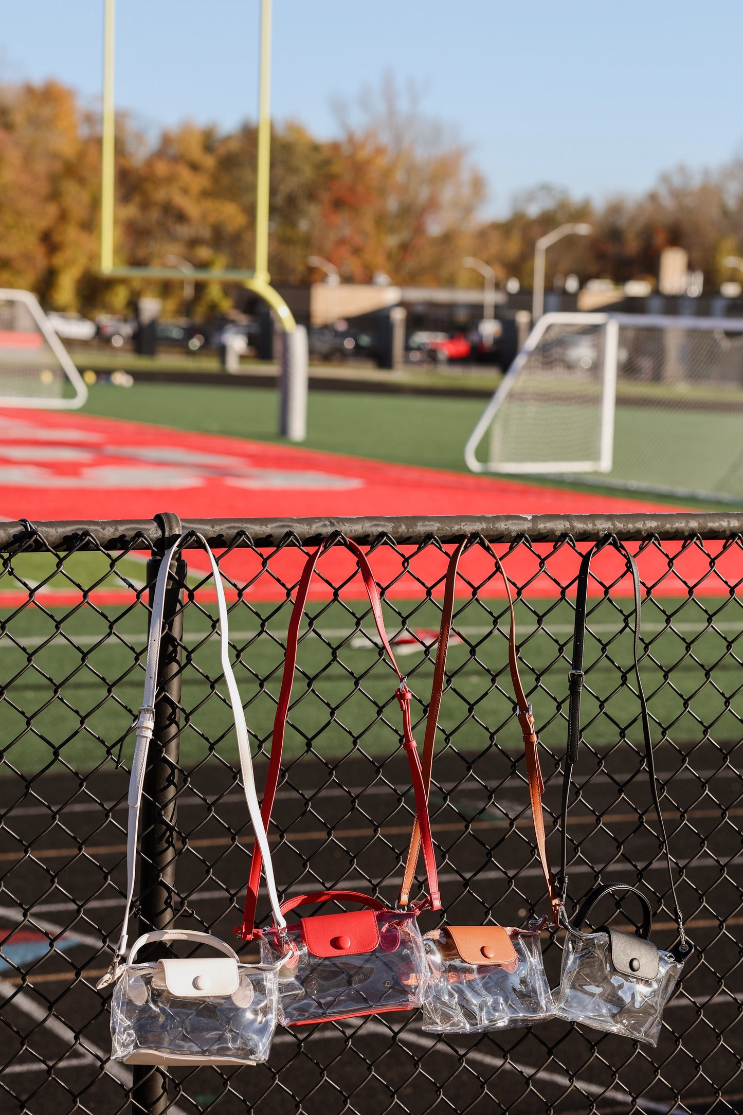 Clear stadium bags with handles attached to a chain-link fence on a sports field.