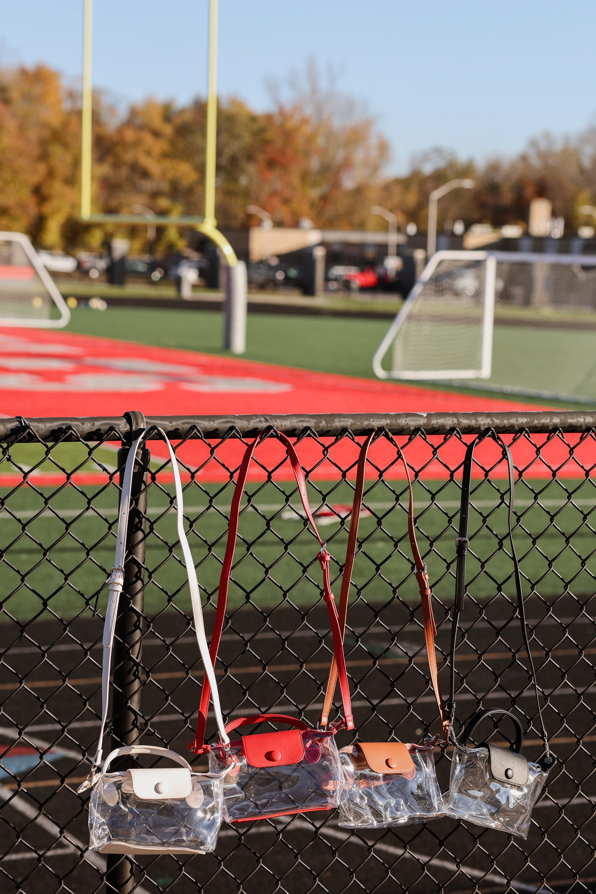 Clear stadium bags with handles attached to a chain-link fence on a sports field.