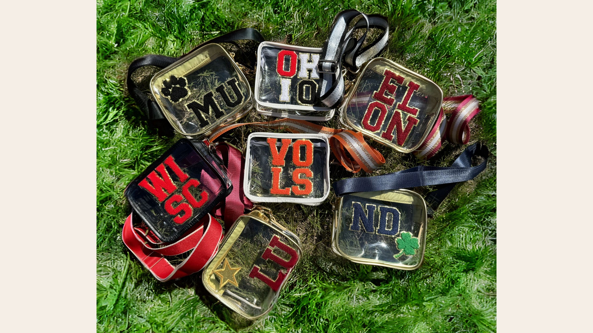 A collection of various clear stadium purses with chenille patches laying in the grass