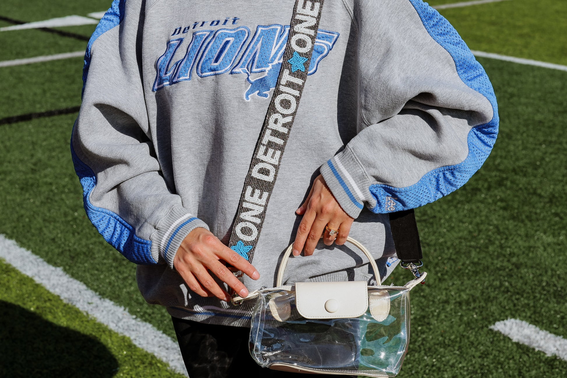 Person wearing a Detroit Lions hoodie holding a clear handbag  with a Detroit lions beaded purse strap on a sports field.