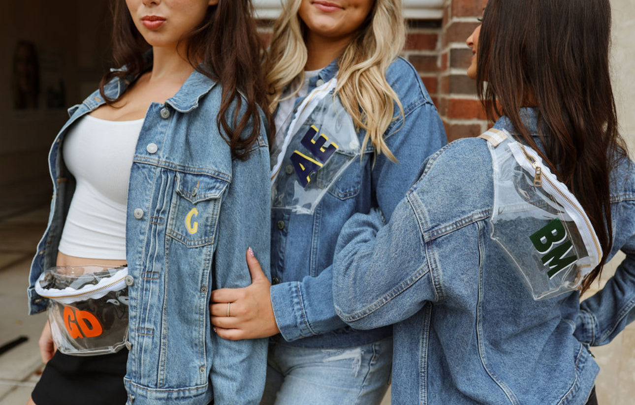 Three women wearing denim jackets with patches and clear monogrammed fanny packs standing close together.