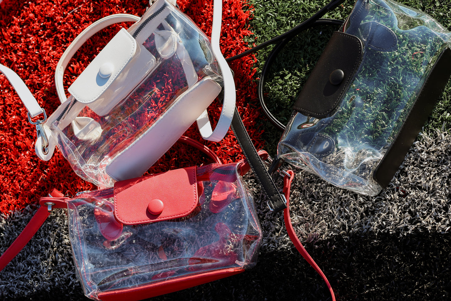 Three clear stadium handbags with leather detail in red, white and black laying on a football field