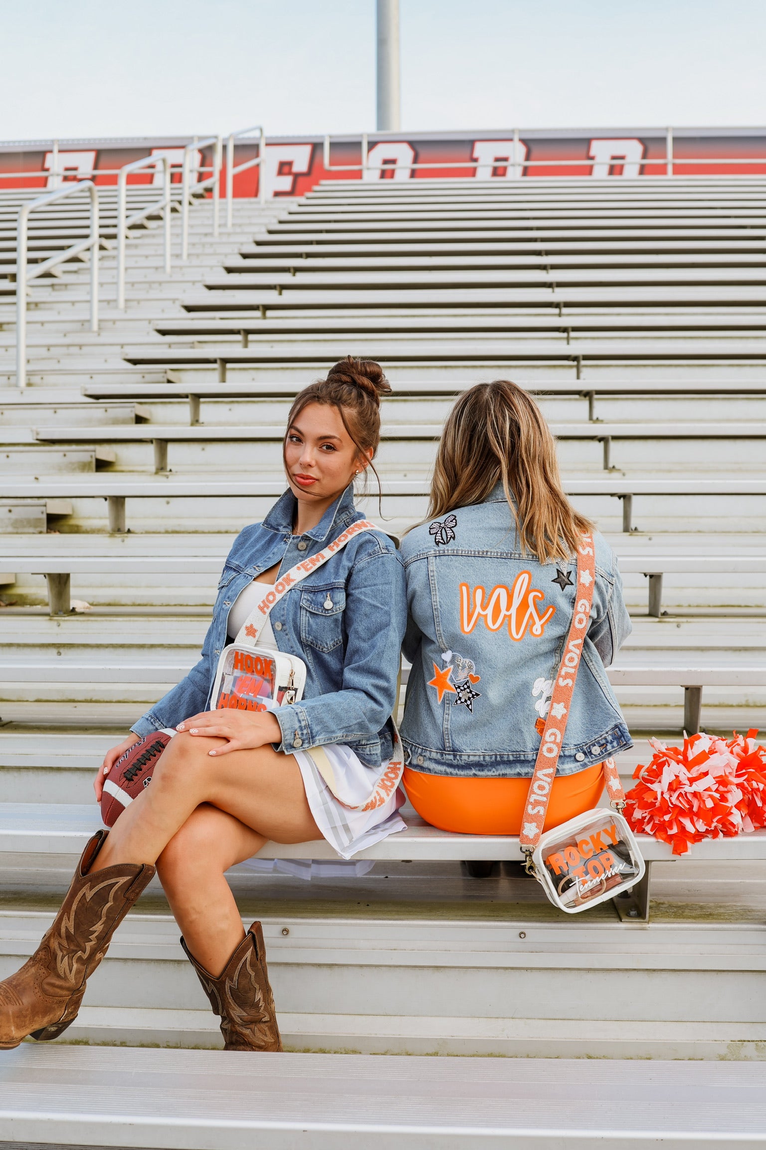 Two women sitting on bleachers with denim jackets and orange accents with clear stadium purses and beaded game day straps